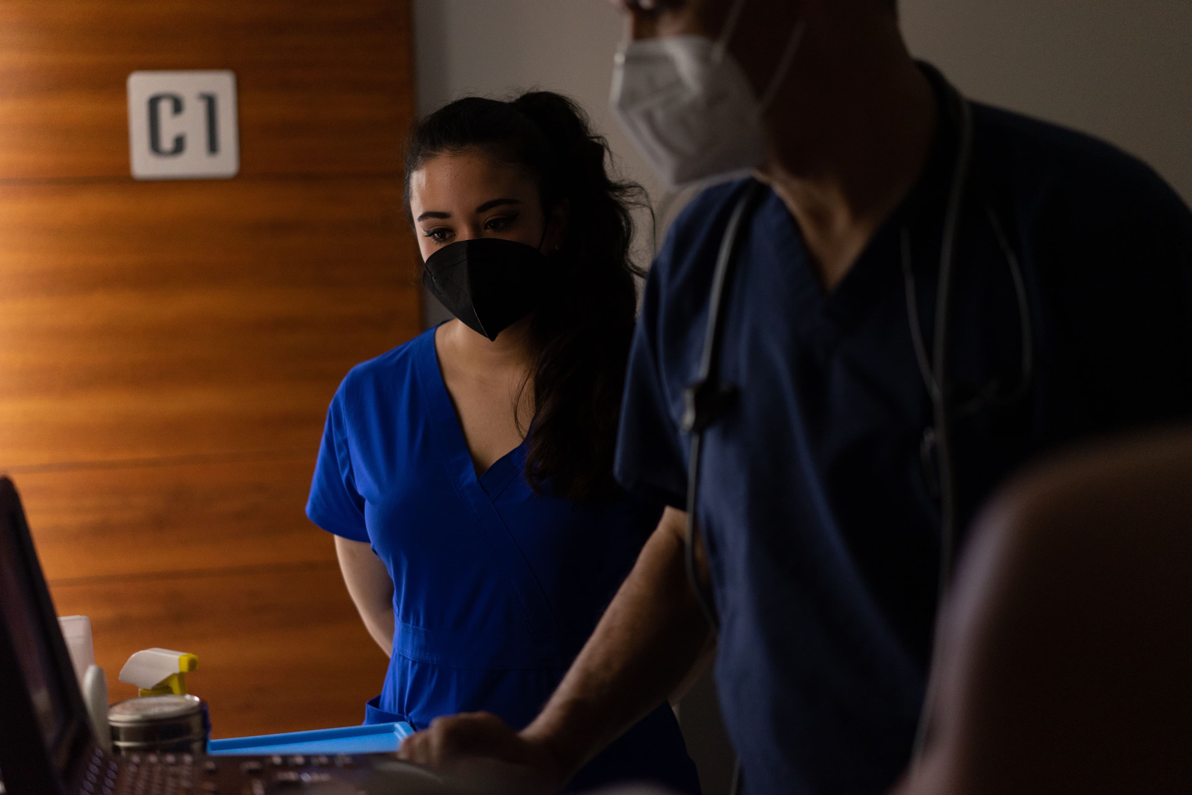 Two masked healthcare workers review patient information in a hospital room.