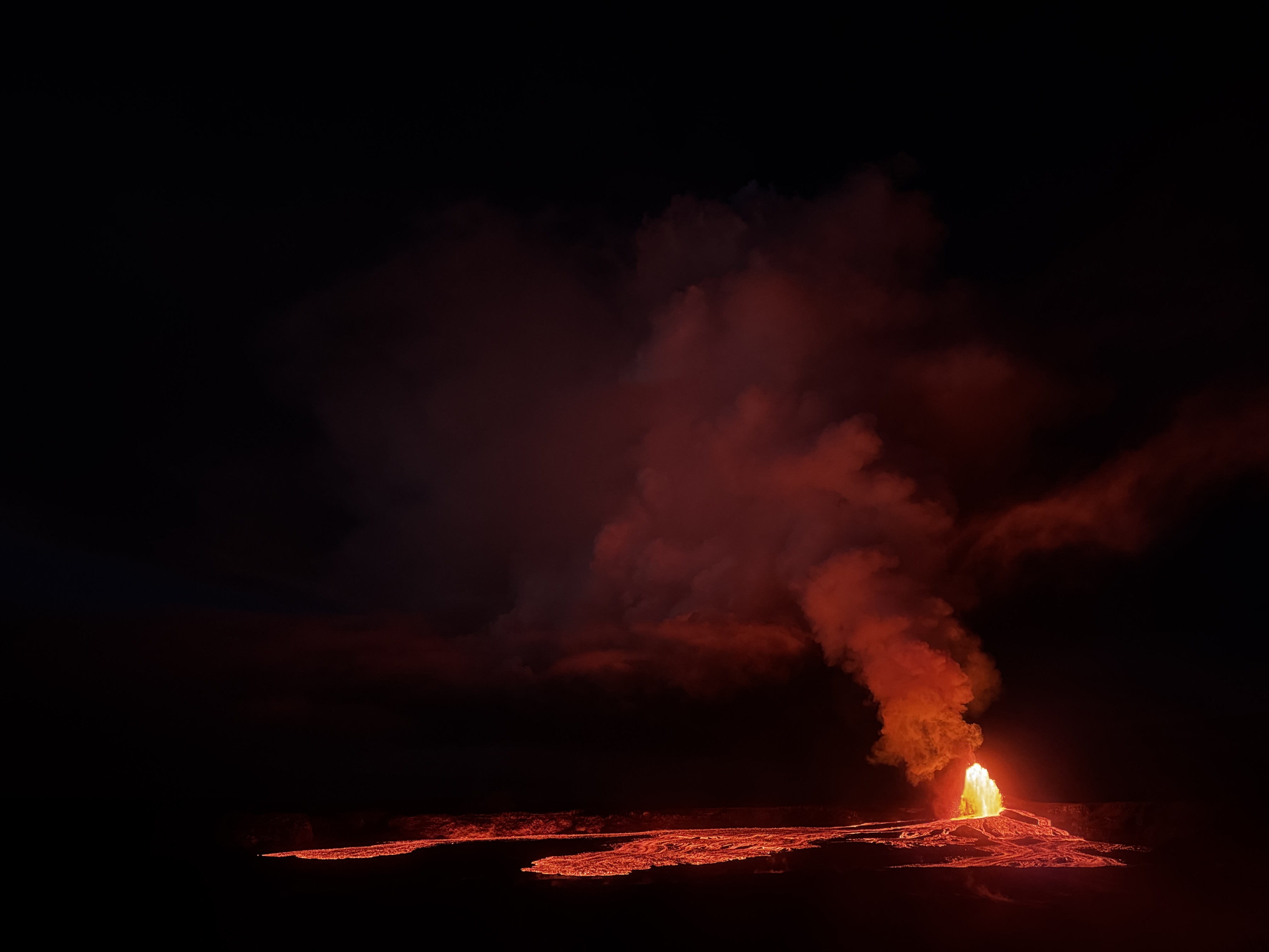 A lava fountain illuminates a plume above glowing lava flows in Halemaumau crater at night.